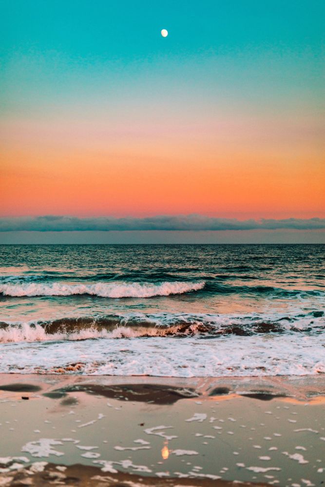 This photo by Ryan Loughlin shows a colorful sunset over a calm ocean with gentle waves rolling onto the sandy beach, pastel sky blending orange and turquoise hues, and the moon visible above the horizon.