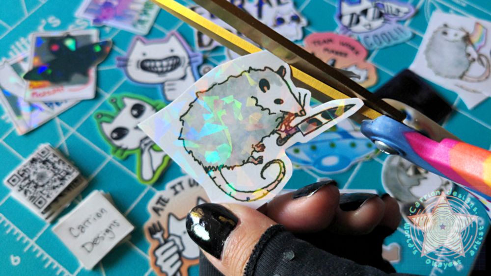 a hand holds an opossum with a knife sticker while cutting the border out with rainbow scissors. an array of similarly quirky and fun cat and cryptid stickers are strewn in the background.