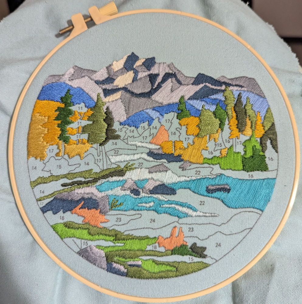 Embroidery hoop in progress depicting a mountain in the background with a forest and river in the foreground.