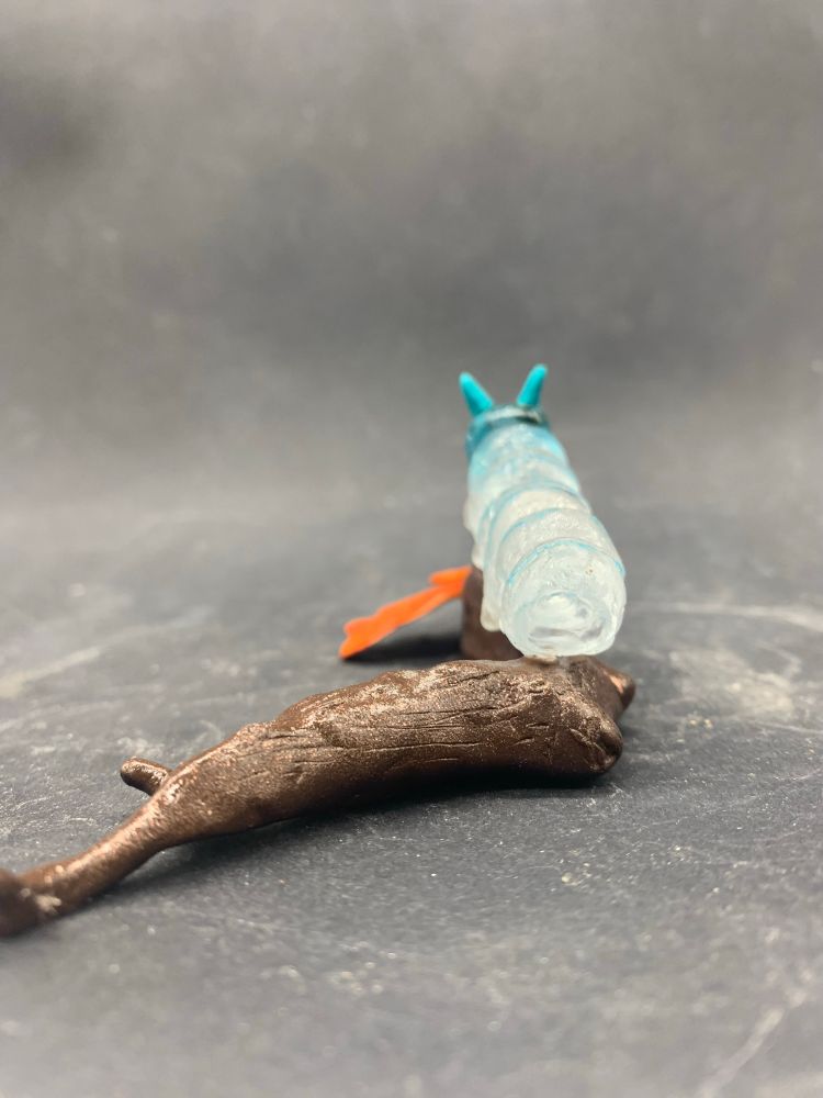 Photo d’une sculpture de chenille en verre transparent et bleu, sur une branche avec deux feuilles orange en argile epoxy, vu de côté vu de derrière 
