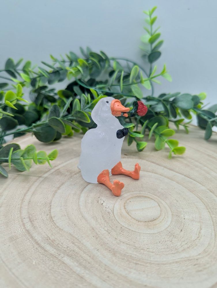 Photo d’une sculpture d’oie assise en cristal balance et argile epoxy, elle a un noeud papillon et un rose dans le bec 
En fond on voit une fausse plante