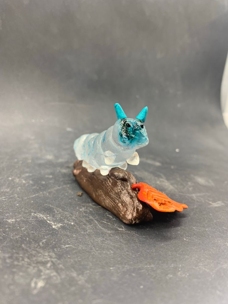 Photo d’une sculpture de chenille en verre transparent et bleu, sur une branche avec deux feuilles orange en argile epoxy, vu de face