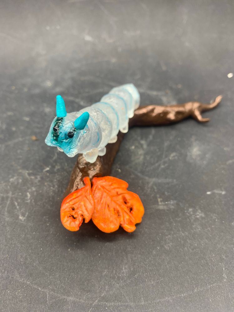 Photo d’une sculpture de chenille en verre transparent et bleu, sur une branche avec deux feuilles orange en argile epoxy