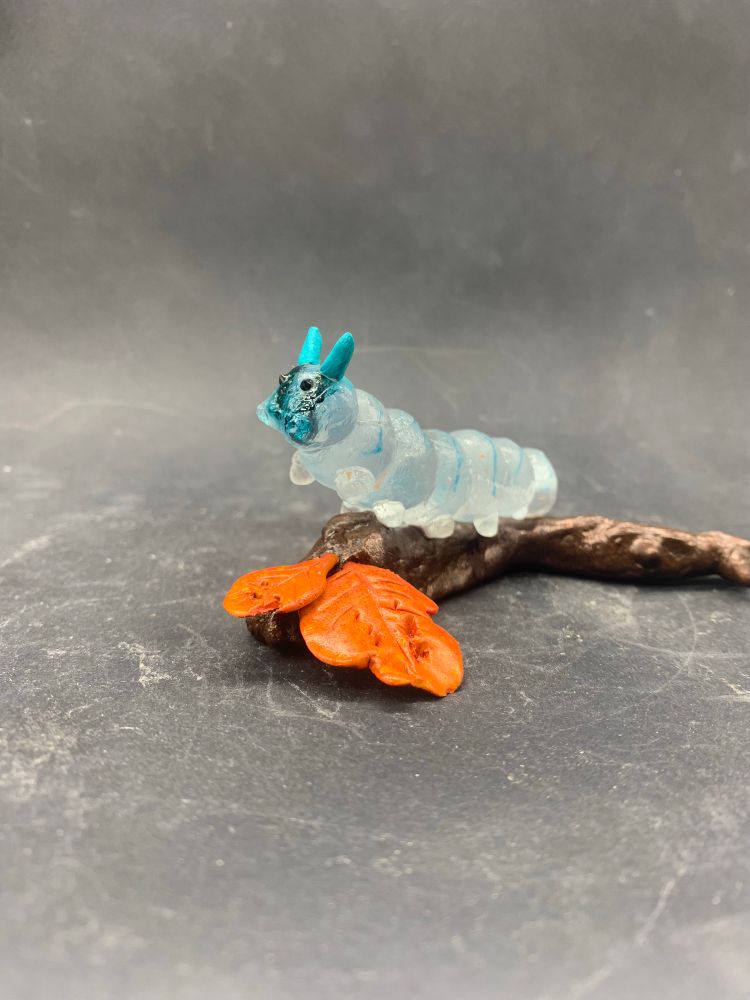 Photo d’une sculpture de chenille en verre transparent et bleu, sur une branche avec deux feuilles orange en argile epoxy, vu de côté 