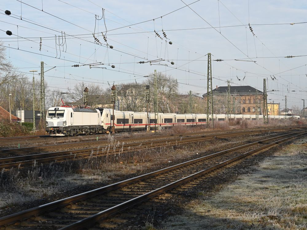 Die Fotografie zeigt einen ICE-L Wagenpark, der von einer weißen Siemens Vectron-Lokomotive gezogen wird. Er fährt auf einer mehrgleisigen Bahnstrecke, die mit Oberleitungen überspannt ist. Im Hintergrund sind alte Bahnhofsgebäude und einige Bäume zu sehen. Der Boden ist mit Frost bedeckt.