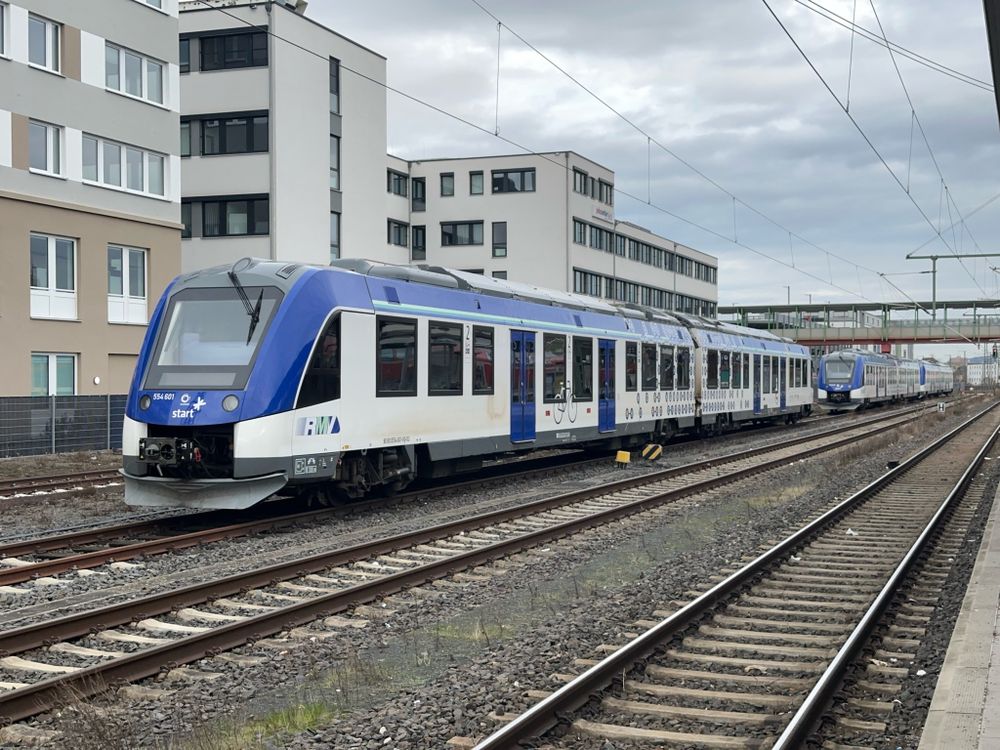 Mehrere moderne weiß-blaue Dieseltriebwagen des Typs Alstom Coradia LINT, mit der Aufschrift „RMV“ und „start“, steht an einem Gleis in einem Bahnhof. Im Hintergrund befinden sich mehrstöckige Gebäude und eine Überführung.