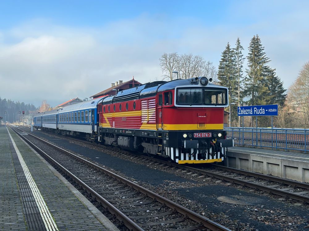 Eine rot-gelb-schwarze Diesellokomotive mit der Nummer 754 074-3 steht mit blauen Reisezugwagen an einem Bahnsteig im Bahnhof Železná Ruda - Alžbětín. Im Hintergrund sind das Bahnhofsgebäude, Bäume und ein leicht bewölkter, blauer Himmel zu sehen.