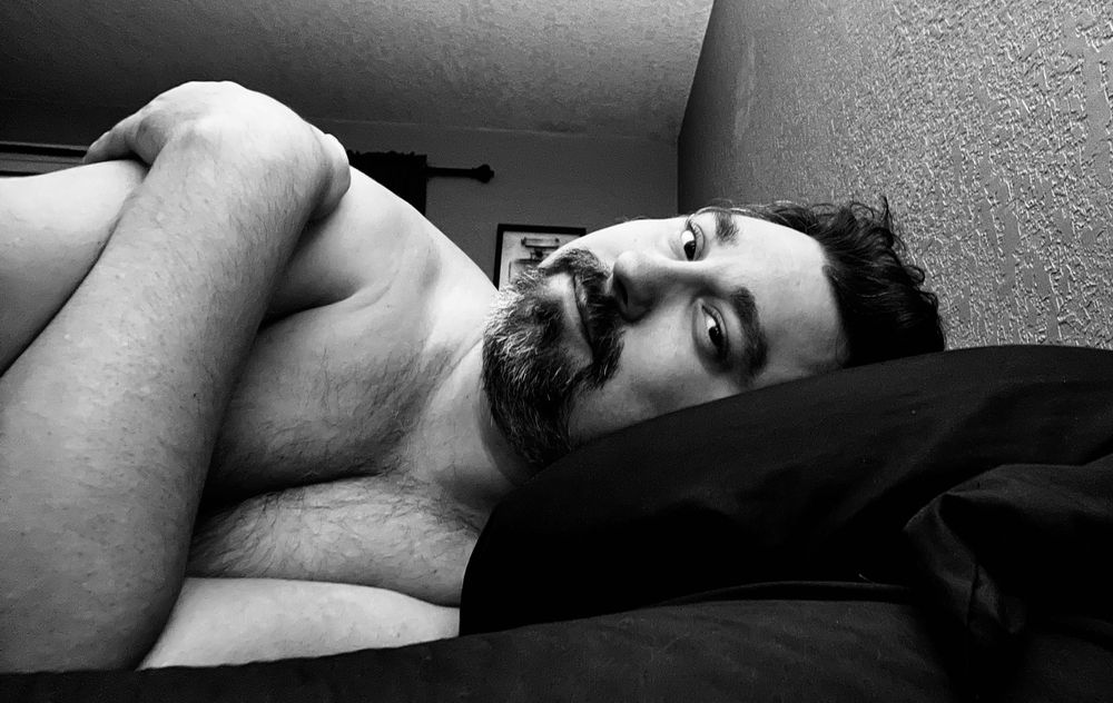 A black and white picture of a shirtless man lying in bed on his side facing the camera 