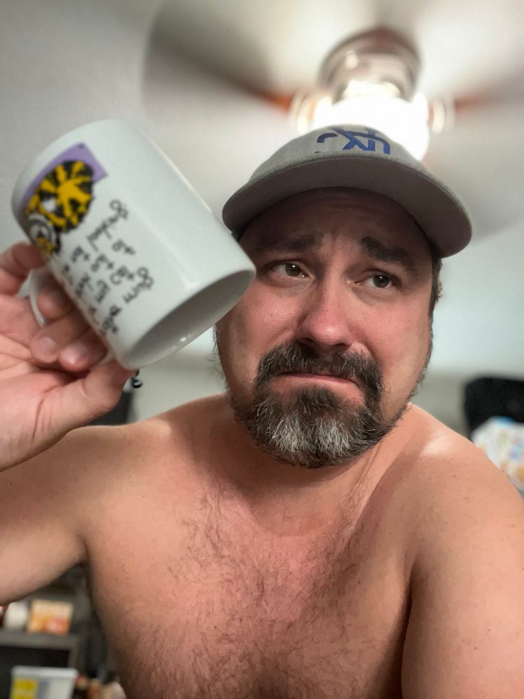 A shirtless man holding an empty coffee mug upside down, looking sad.