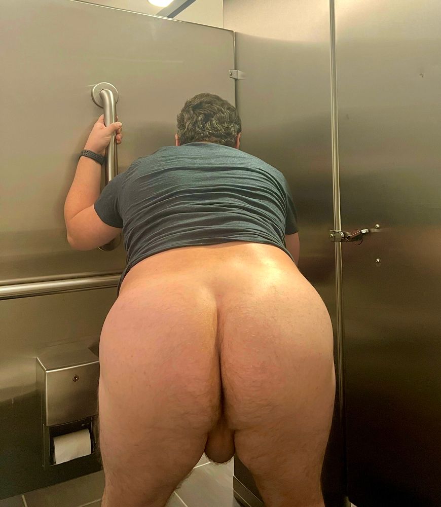 A man with his pants down showing his ass and balls, leaning against the side of a bathroom stall.