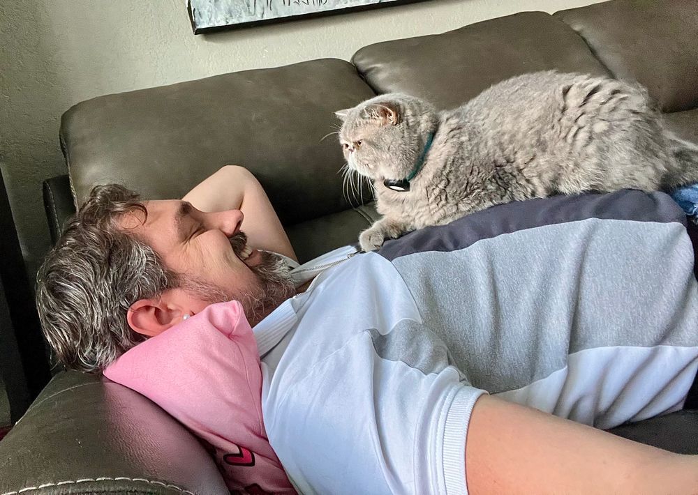 A man lying on a couch with a fluffy kitty on his belly, both looking at each other