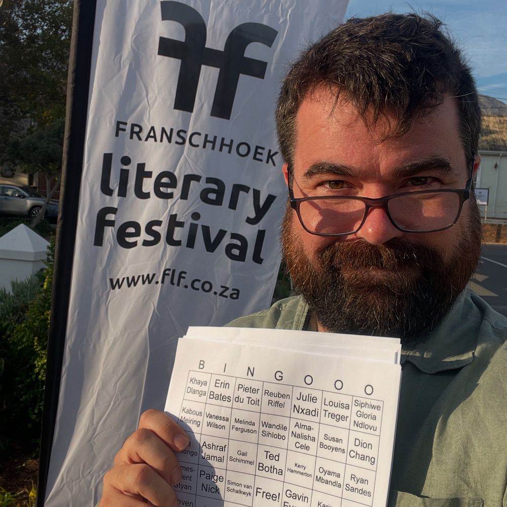 Sam Wilson at the Franschhoek Literary Festival holding a stack of homemade author bingo cards 