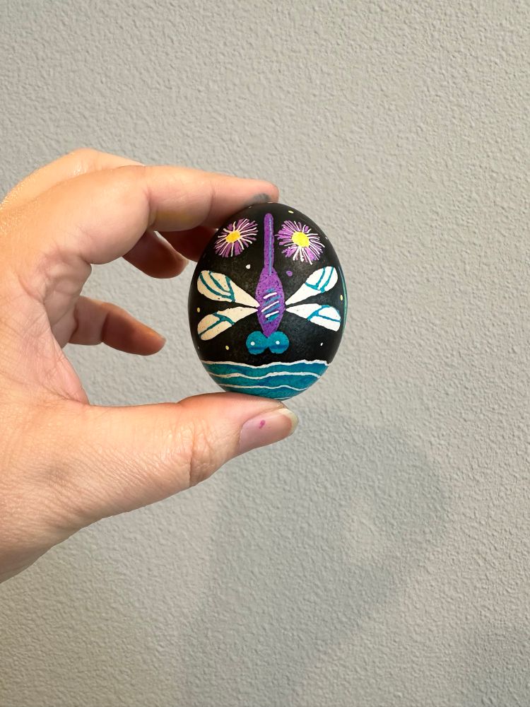 A hand holding a decorated easter egg (pysanky) vertically in front of a light grey wall.  The egg is black, with a purple background dragonfly with blue eyes in the center.  On the top half of the egg, to either side of the dragonfly is a pink and white english daisy with a yellow center.  Under the  dragonfly are blue and white water ripples.