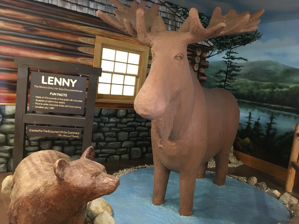 A lifesize chocolate moose standing in a blue pond near a cabin facade. That’s moose with two Os. There’s also a smaller bear. The sign declares: 

LENNY
The World's Only Life-Size Chocolate Moose
FUN FACTS
Made of 1700 pounds of fine grade milk chocolate
Sculpted on-site in four weeks
Pond is white chocolate tinted with food coloring
Unveiled July 1, 1997
Created For The Enjoyment Of Our Customers
The Hamond Family