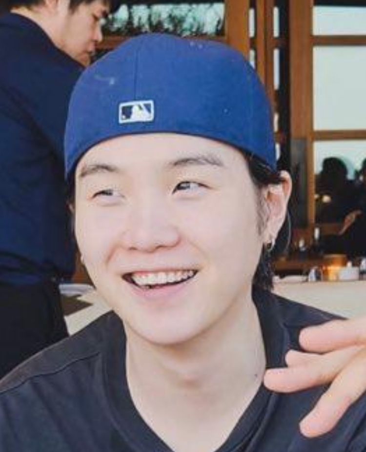 BTS member Yoongi smiling; it’s a closeup shot, from the shoulders up, and he’s SMILING; he’s wearing a dark t-shirt and a blue baseball cap backwards and he’s SMILING YOU GUYS; he appears to be at a restaurant based on the background but no one’s looking at the background when he’s SMILING LIKE THAT because all that’s important is he’s sitting at a table with his bandmates, his brothers, and smiling so happily YOONGI IS HAPPY EVERYONE 