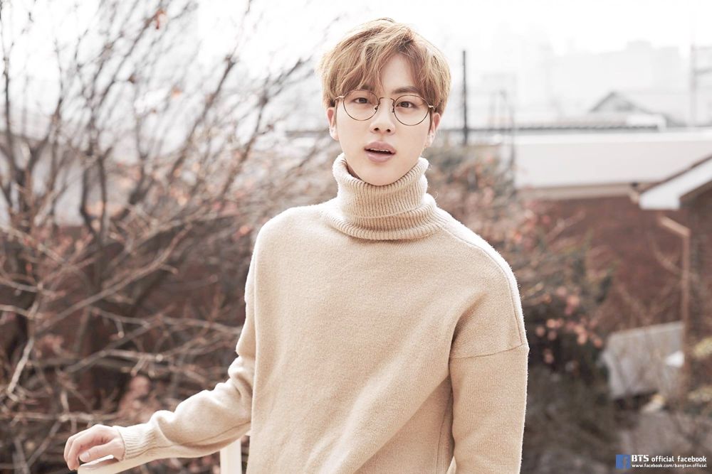 BTS member Jin with caramel-blond hair, standing outside while wearing a camel-colored, really soft-looking turtleneck sweater and circular wire-frame glasses; he is looking directly into the camera with a small open-mouthed smile