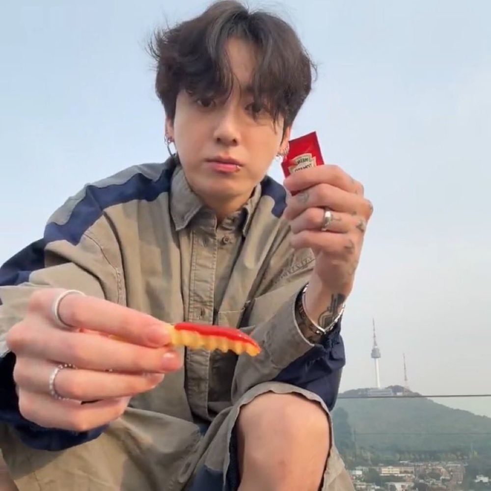 BTS member Jung Kook, live streaming from on top of his roof with the Namsan Tower atop a mountainside behind him, is holding out a french fry so his viewers can see it; he has perfectly placed a line of ketchup across the top of the french fry; he is very good at mukbangs, someone give him a gold star or some stickers or some gold star stickers, he deserves it