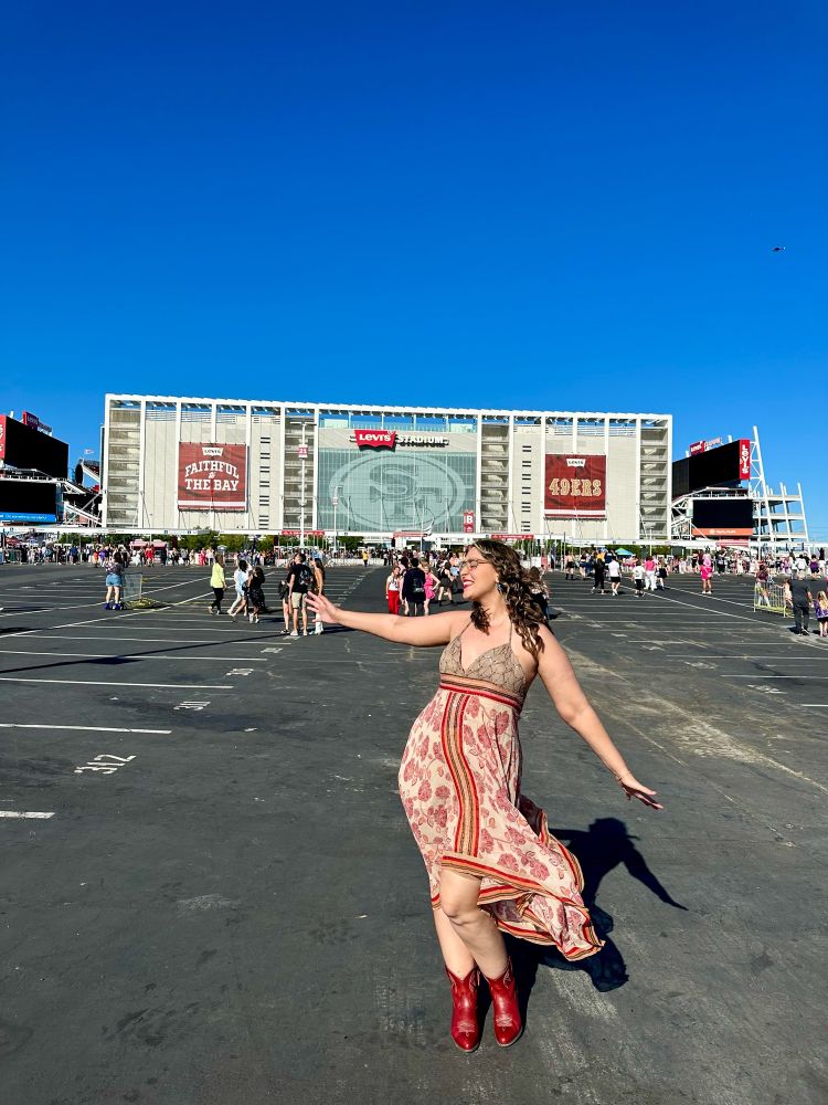 Laura standing in front of Levi Stadium in Santa Clara wearing a red & tan dress with a handkerchief style hem and red cowboy boots. The dress is fluttering in the wind and is the same dress Taylor Swift wore in her debut era. 