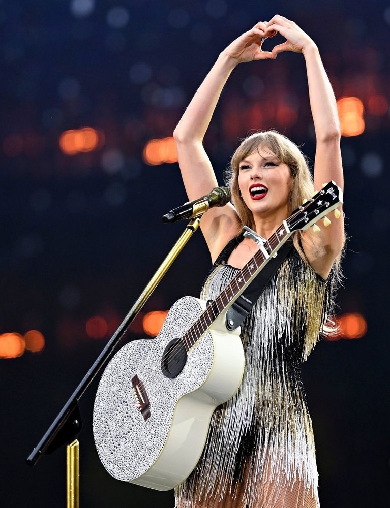 Taylor swift doing heart hands and smiling at the crowd during an eras tour show 