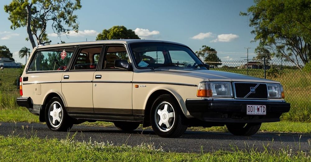 A picture of a cream-colored Volvo 240 wagon next to a grassy field.
