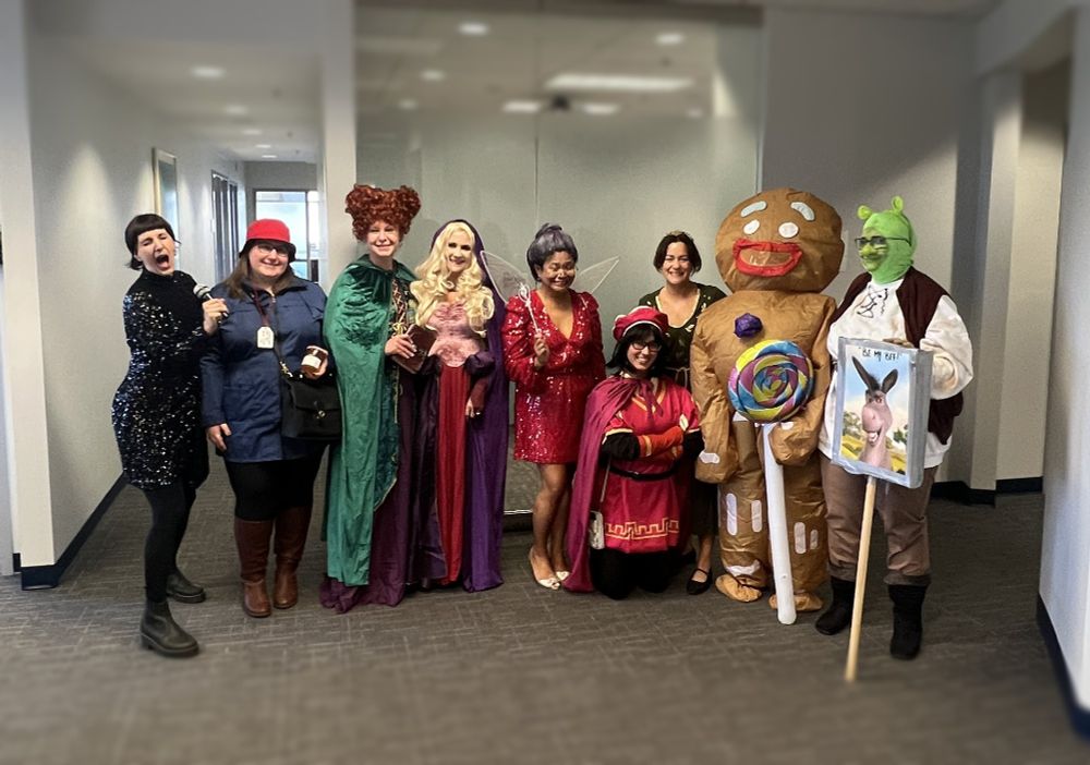 Group of people dressed in various costumes, including characters from Disney and Shrek, and a Gingerbread Man, posing together in an office hallway.