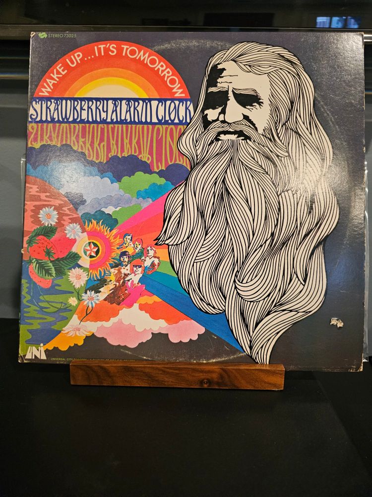 The front cover for Wake Up...It's Tomorrow. Half the cover is an artistic rendering of the band in a colorful cluster of clouds and rainbows. The other half is a black and white drawing of an old man with long hair and a long beard.