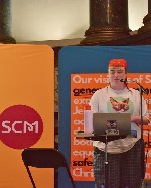 One of the speakers at National Gathering, Kate Bradley (they/them) standing in front of an SCM banner while they gave their talk.