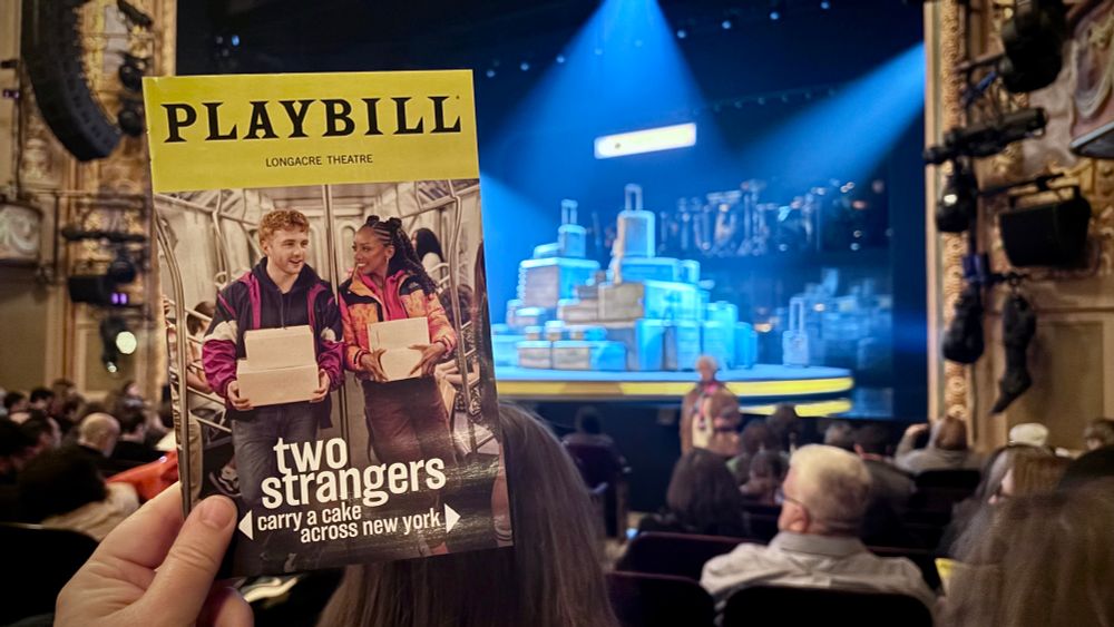 Playbill held up in front of the set, a collection of rotating luggage. 