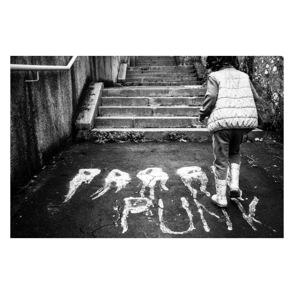 A young child, seen from the back and slightly to the right, stands at the bottom of a set of weathered concrete stairs outdoors. They are wearing a light-colored, quilted, puffy vest over darker clothing and white boots. The ground before the stairs is dark asphalt, cracked and uneven. Scrawled in large, rough, white letters on the ground are the words "PARAF PUNK". The overall image is in black and white, lending a gritty, somewhat melancholic or defiant mood. The scene evokes a sense of urban decay juxtaposed with youthful presence.