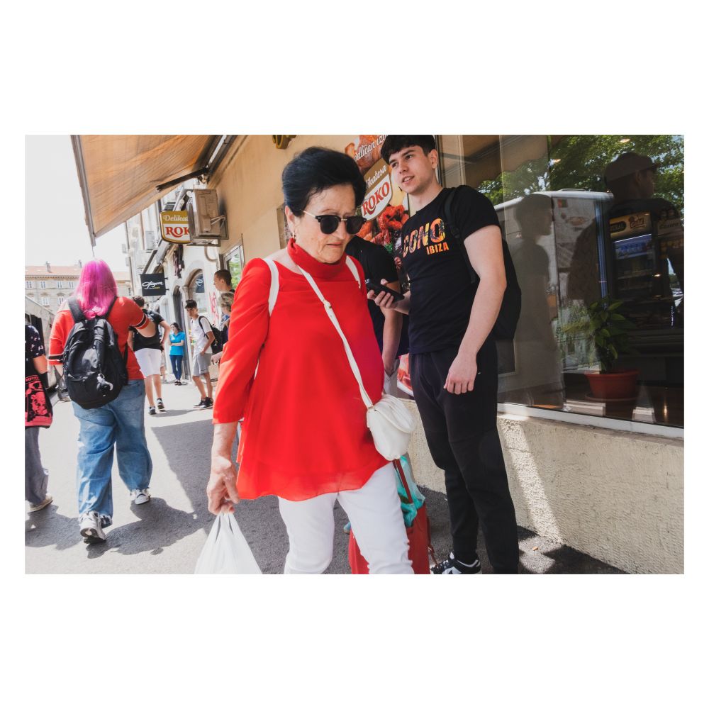 A candid street photograph shows a woman in a vibrant red top and white pants walking towards the viewer, wearing sunglasses and carrying a white shoulder bag and a white shopping bag. To her right, a young man in a black t-shirt stands near a storefront. Other pedestrians and shop facades are visible in the background under bright sunlight.