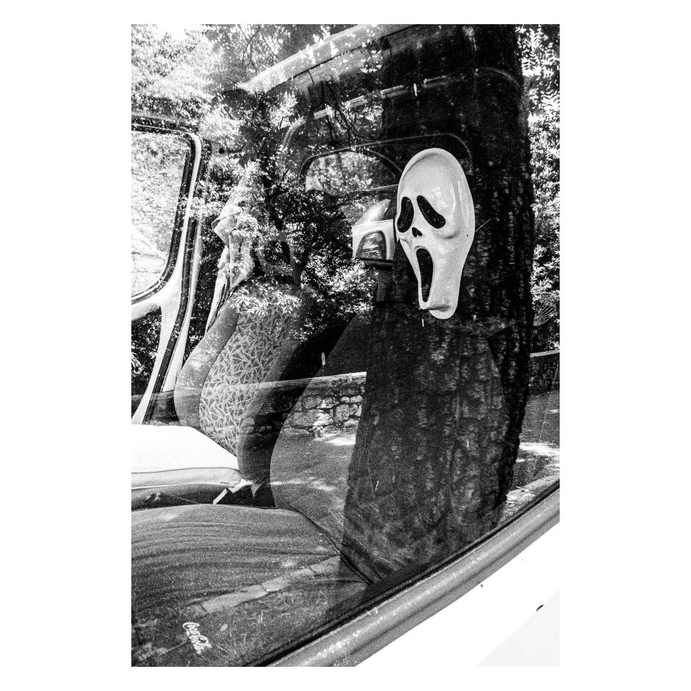 A high-contrast, black and white photograph creates a scary movie aesthetic. Inside a car, a Ghostface mask is positioned on the passenger seat, its mouth open in a silent scream. The reflection of dense trees on the car window adds to the eerie, unsettling mood.