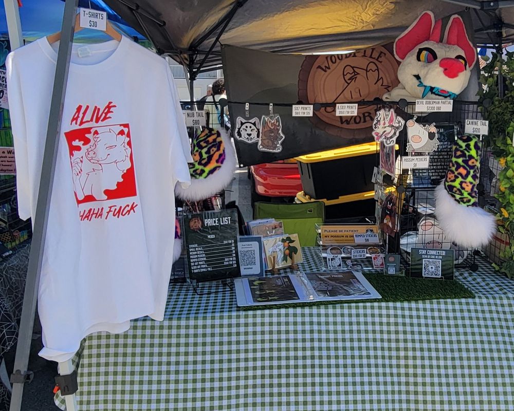 My vending display. From left to right, a tshirt with the "Alive Ahaha Fuck" design (featuring a possum smoking a blunt), one of the premade nub style tails,  badges hanging from the grid display,  art prints, stickers, the doberman premade,  and another premade canine style tail. In the background is my company sign hanging from the canopy.  Not pictured is the sound of sizzling meat as we roasted under the heat of the sun. 