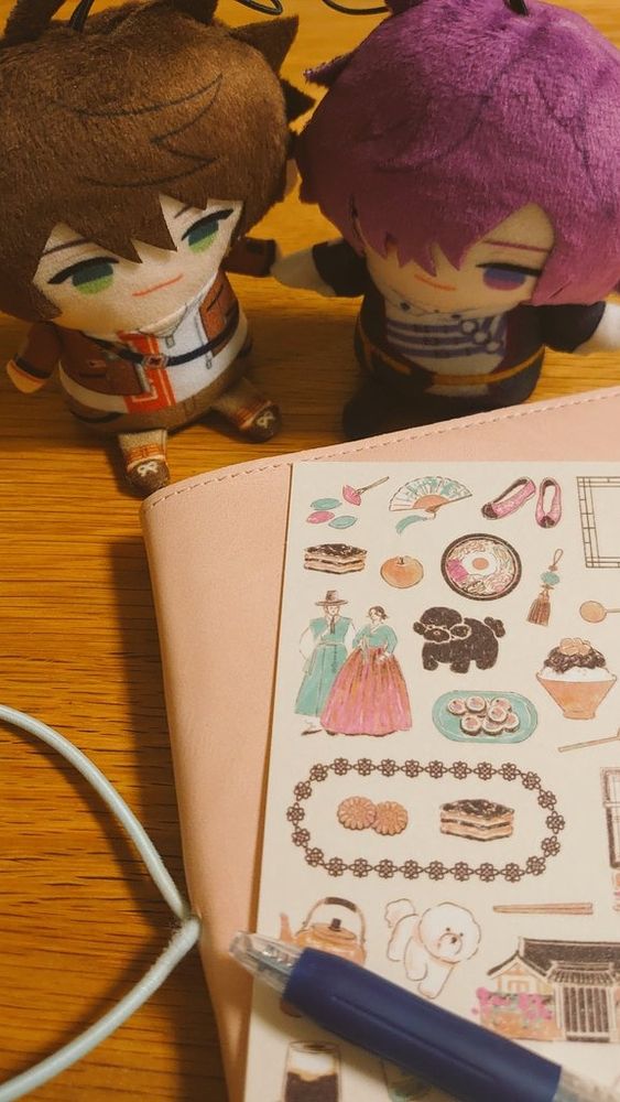 木製テーブルの上にスハと浮奇のにじパペとピンクのカバーの手帳が置かれている。手帳の上には韓国文化をモチーフにしたステッカーとペンが1本置かれている。