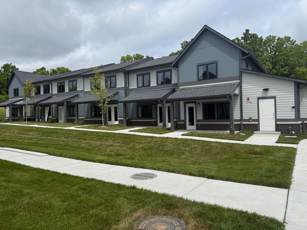 This image captures a row of newly constructed, modern townhouses featuring a cohesive design aesthetic under an overcast sky. The buildings are two stories, with a blend of dark gray and slate blue siding complemented by white trim around the windows and along the roofline. Each unit has a covered entrance, and several units display a small, inviting porch area. The townhouses are aligned in a straight row, showcasing symmetry and uniform landscaping. The fresh, manicured green lawn in front is divided by new concrete walkways that lead to each front door, contributing to the clean and orderly appearance of the neighborhood. Notably, there’s a drainage grate embedded in the walkway and a fire department connection (FDC) prominently displayed on one end of the building, indicating attention to safety and accessibility in the development's planning. The setting is serene, bordered by a line of young, planted trees and a backdrop of lush, mature trees behind the properties, suggesting a blend of urban and natural elements. This image portrays a modern housing development designed for aesthetic appeal and functional living.
