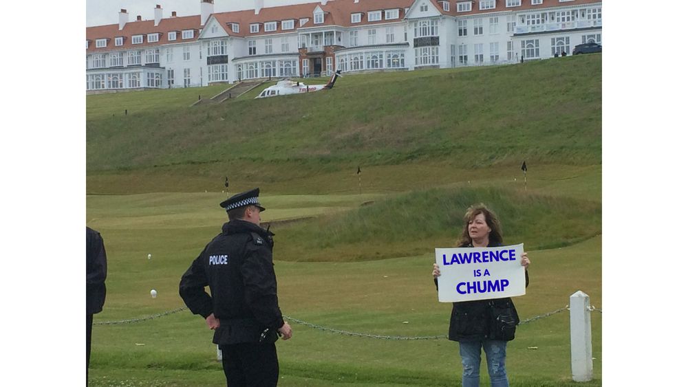The iconic image of the late, great Janey Godley being approached by a policeman as she holds up her famous sign on Donald Trump’s golf course in Scotland. Her sign read ‘Trump is a C***.’ But it has been adapted in this image to read ‘Lawrence is a Chump.’
