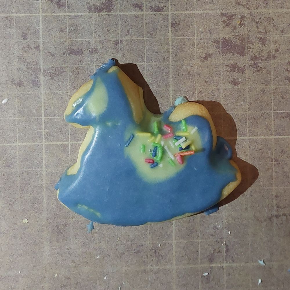 Foto eines weiteren selbstgebackenen Schaukelpferd-Plätzchens, das von mir sehr grob mit blauem und gelbem Zuckerguss und bunten Streuseln dekoriert wurde. Es sieht aus wie ein zerstörtes Lapras, das 15 Kriege erlebt hat.