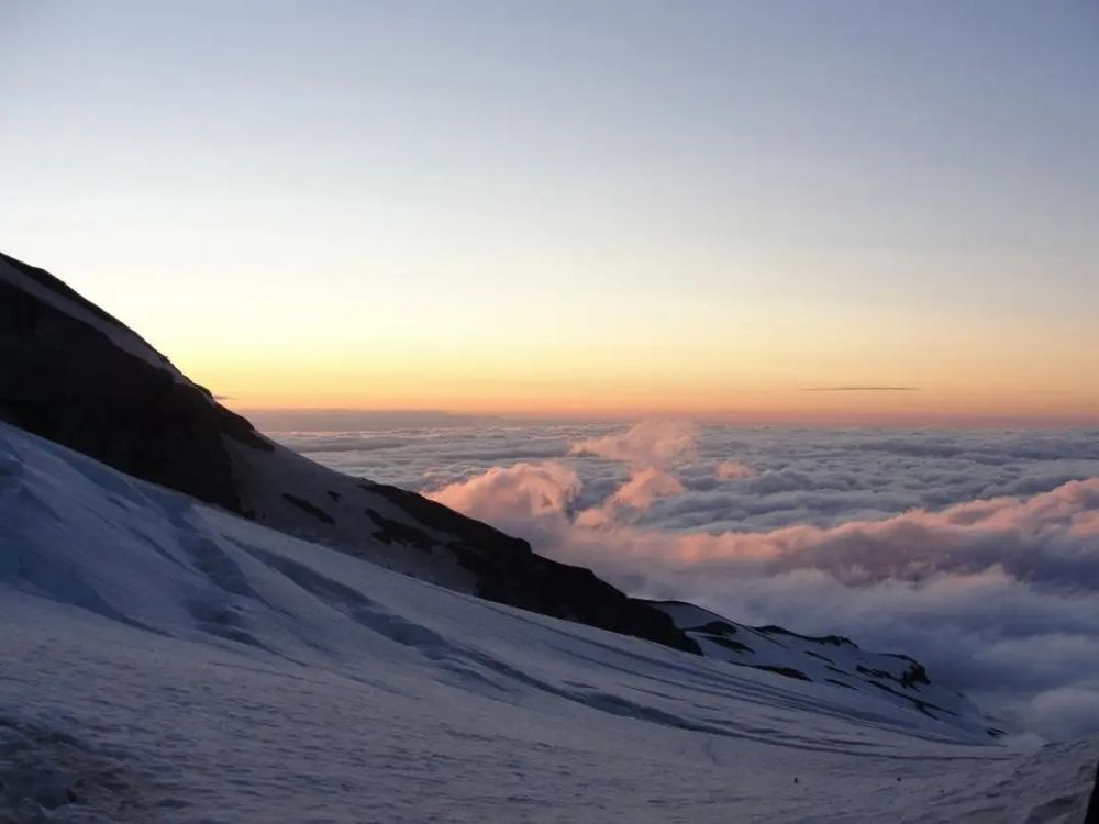 Below the glacier covered slope of the northern face of Mt. Rainier, a sea of seemingly endless clouds stretches into the sunset. The sunset is muted gold and pink, with a pale blue sky above. 
