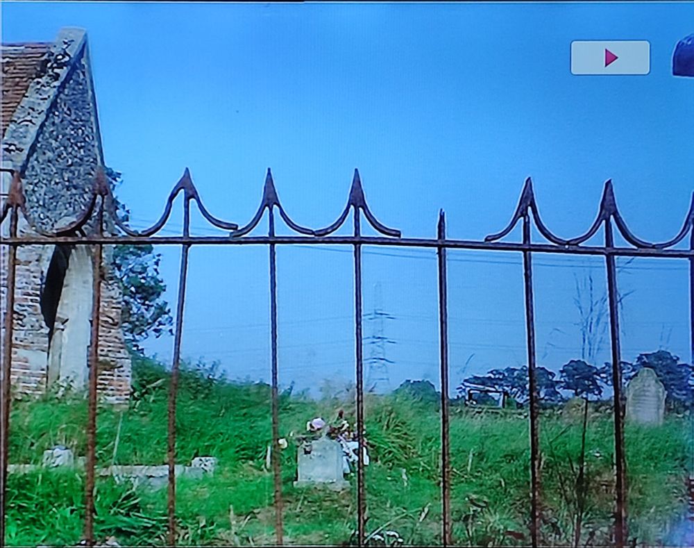 Rusty metal fence with pointy decorative bits on top, in front of a disused church and it's yard. A pylon is visible in the distance 