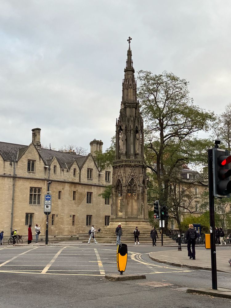 Large slender  gothic cross in front of of 3 storey college with road crossing in foreground 