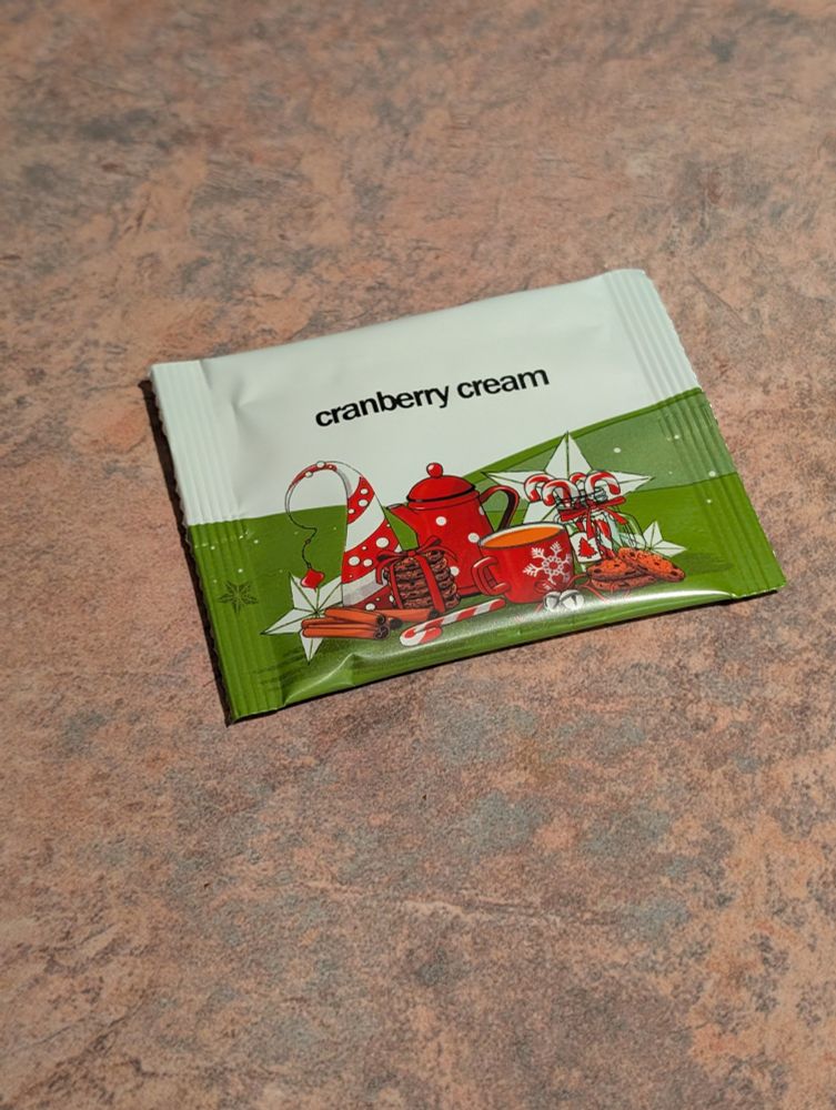 Tea packet labelled cranberry cream. Packet is white and green with a picture of a red tea pot, mug, cookies, candy canes.