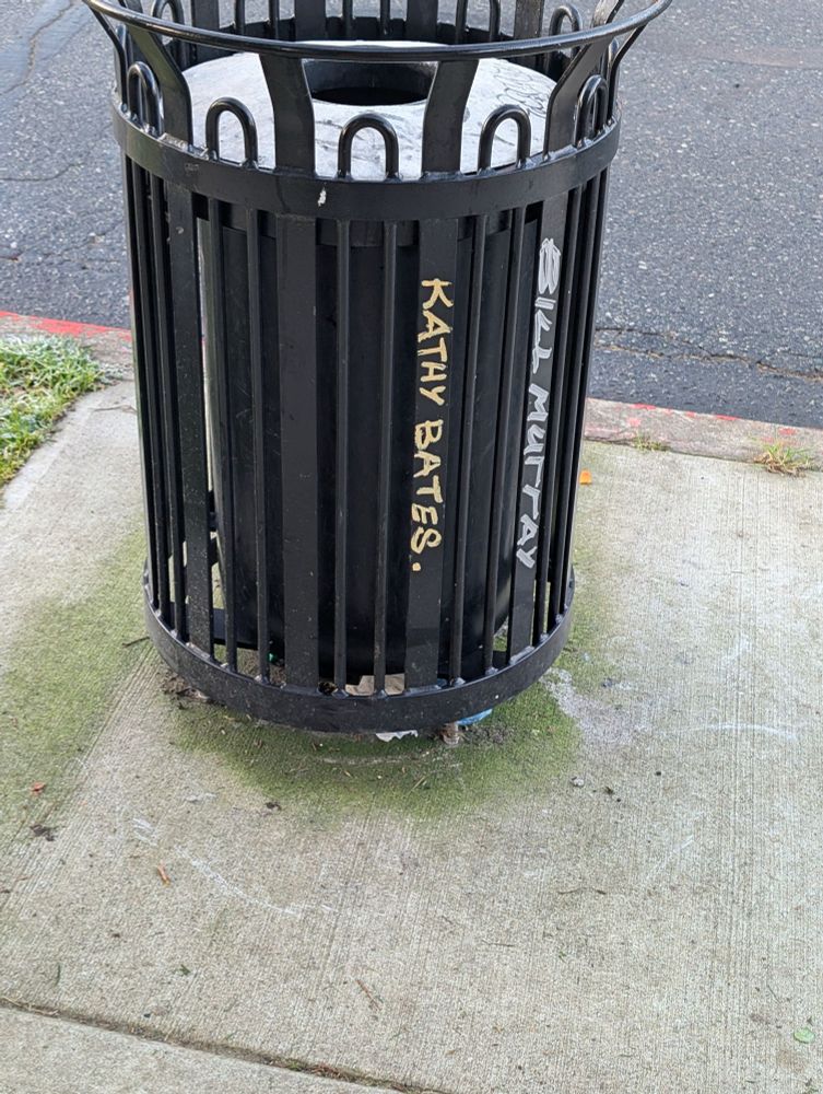 A black metal street garbage bin with graffiti "Kathy Bates" and "Bill Murray"