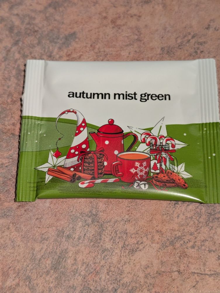 A green and white tea packet with red and white images of a mug, a tea pot, a stack of cookies, candy canes and more