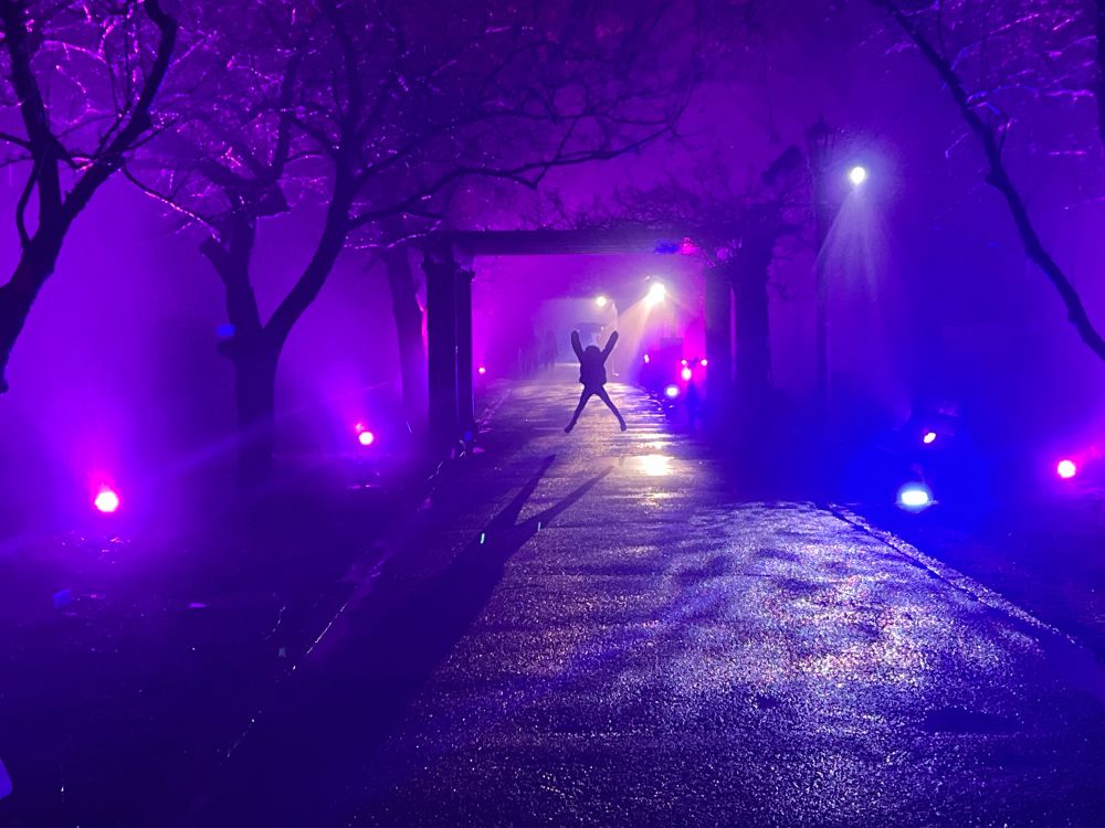 A small child is silhouetted jumping in the air, on a dark garden pathway lit by purple spotlights. 