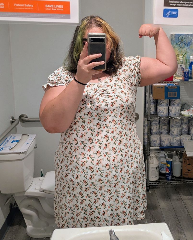 a mirror selfie, i'm wearing a white dress with red and pink flowers on it and flexing my bicep