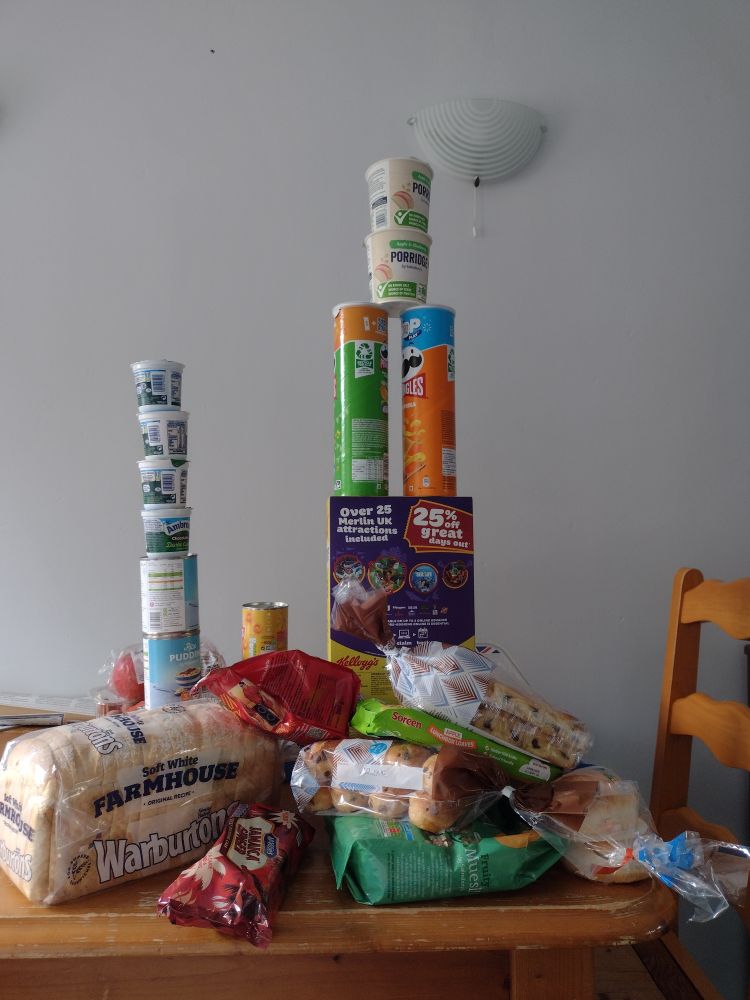 Towers of food on the table - cereal boxes, Pringle tubes, tins of rice pudding and chocolate custard.