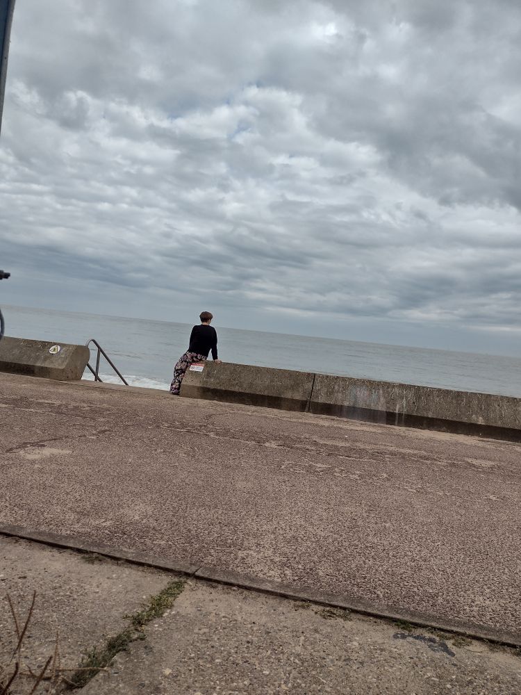 Concrete walkway by the sea. Sea and sky both looking quite grey behind it. In the distance is a woman in a black jumper looking out to sea.