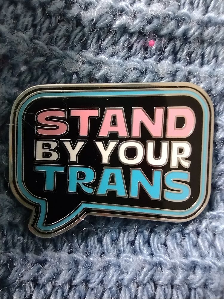 A pin badge which reads Stand by your trans, in pink, white and blue, pinned to my pale blue jumper.