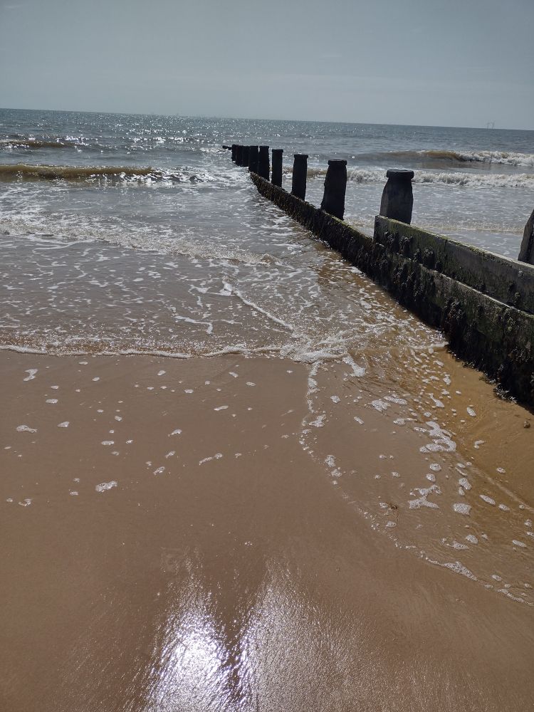 A photo of the beach, sand covering most of the photo, wooden groynes on the right, the sea coming in, and a blue-grey sky above.