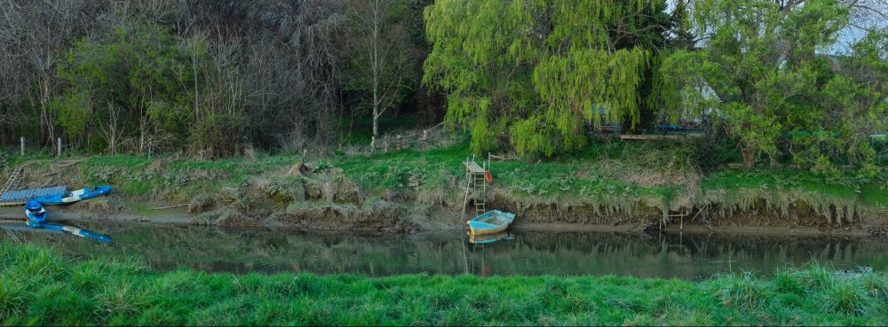 a view across a narrow stretch of river, showing a small boat tied up by the bank, beneath a green willow tree