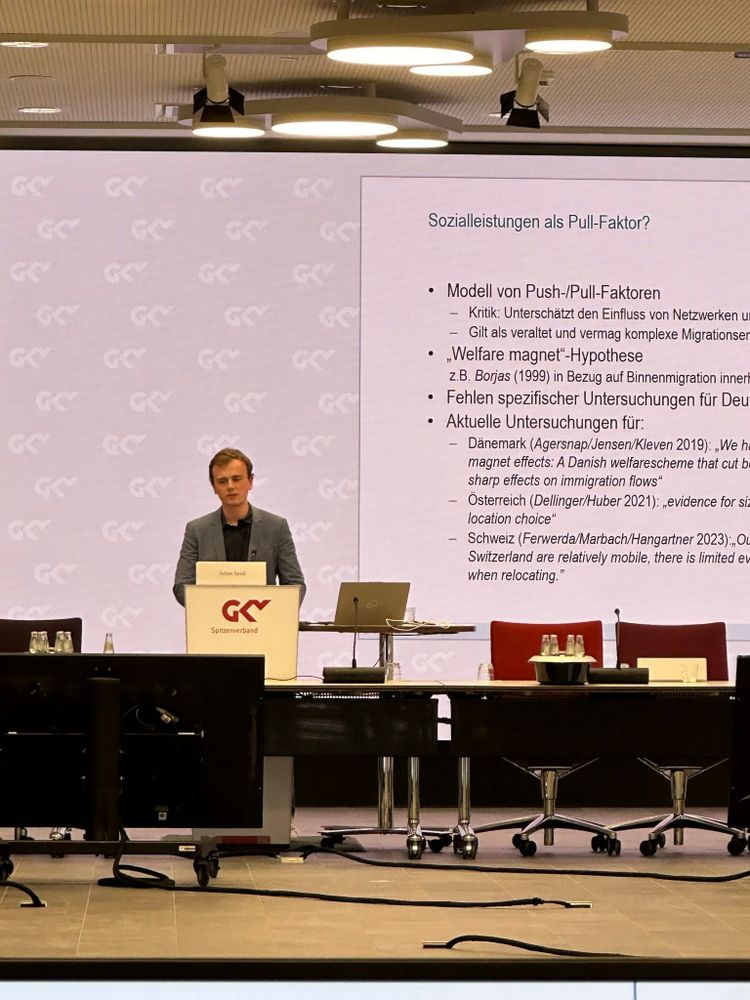 Julian Seidl als Referent am Rednerpult.
Powerpoint-Folien mit der Überschrift "Sozialleistungen als Pull-Faktor" im Hintergrund.